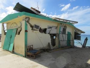 Damaged yellow and blue fishing building stands on cement slab with crystal blue water in background