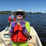 A person donning purple gloves, an orange life vest, sunglasses and a wide-brimmed hat holds a small fish.