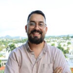 A person with a short dark beard and glasses smiles. An urban landscape is behind him.