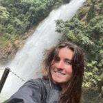 A woman with long brown hair smiles in front of a waterfall.